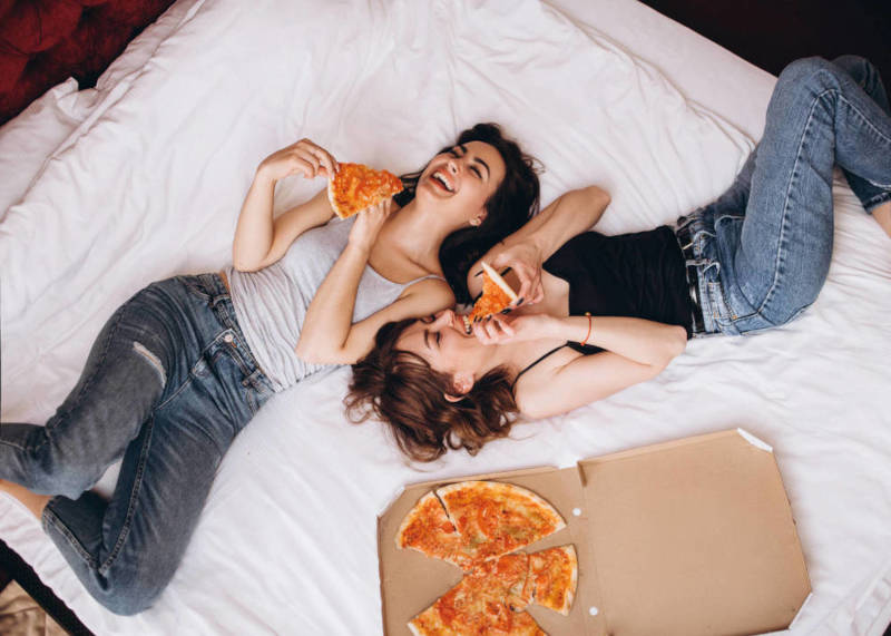 zwei junge Frauen essen Pizza aus Pizzakarton auf Bett liegend